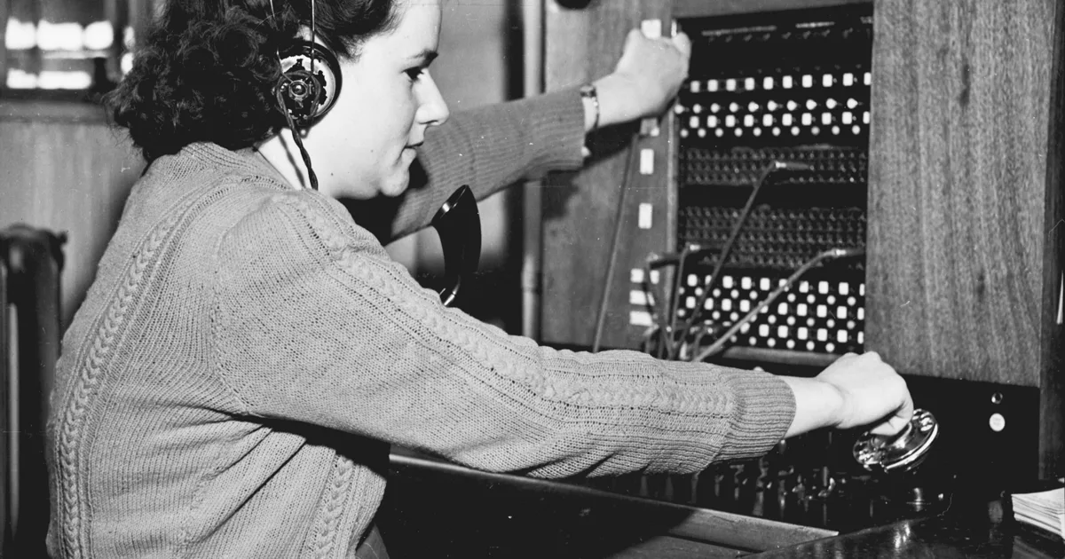 Fotografía en blanco y negro de una operadora con audífonos que conecta llamadas a mano en un conmutador telefónico de clavijas, en una oficina de 1949